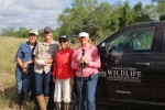 Four graduate students in outdoor field gear stand beside a black pickup truck labeled “Caesar Kleberg Wildlife Research Institute, Texas A&M University–Kingsville.” One person holds a drone controller, and another holds a field tool, with tall grass and trees in the background under a partly cloudy sky.